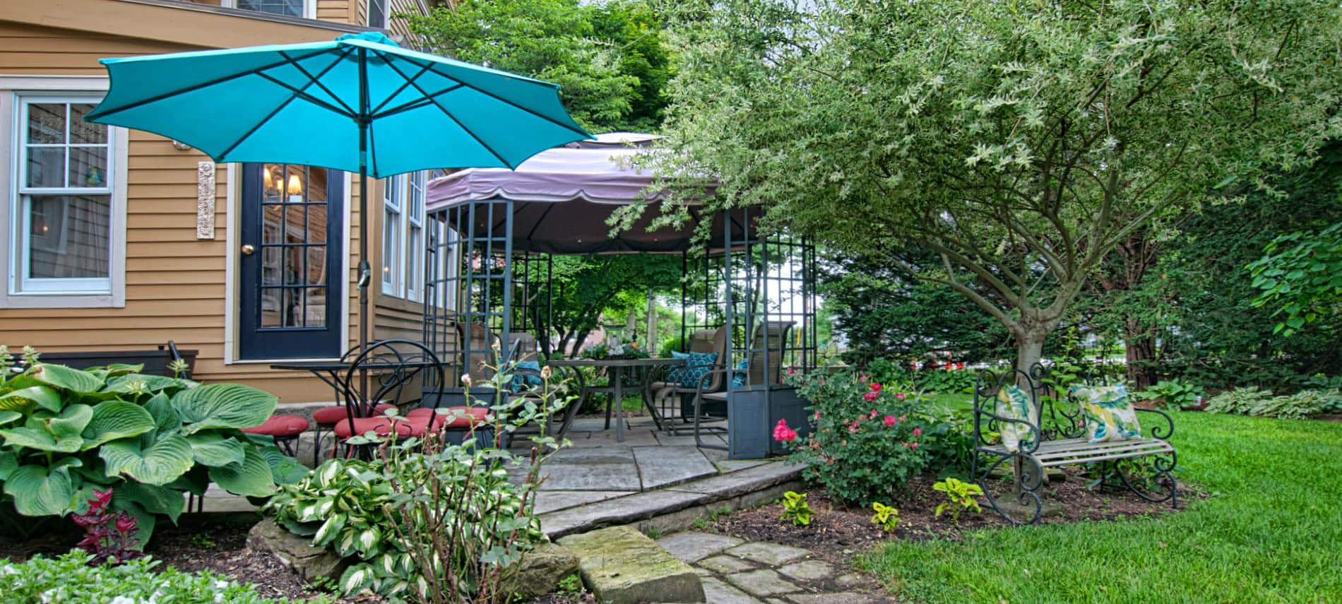 Rear patio with umbrella tables and chairs surrounded by lush green grass, plants and trees