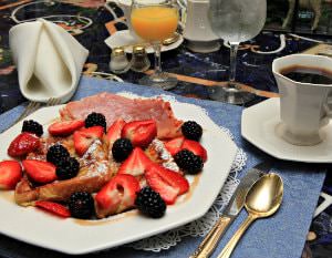 White breakfast plate topped with French toast, fresh strawberries and blackberries, and ham, and orange juice, water and coffee