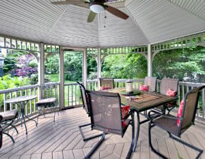 Screened-in gazebo with wood floor, tables and chairs and a ceiling fan surrounded by lush landscaping