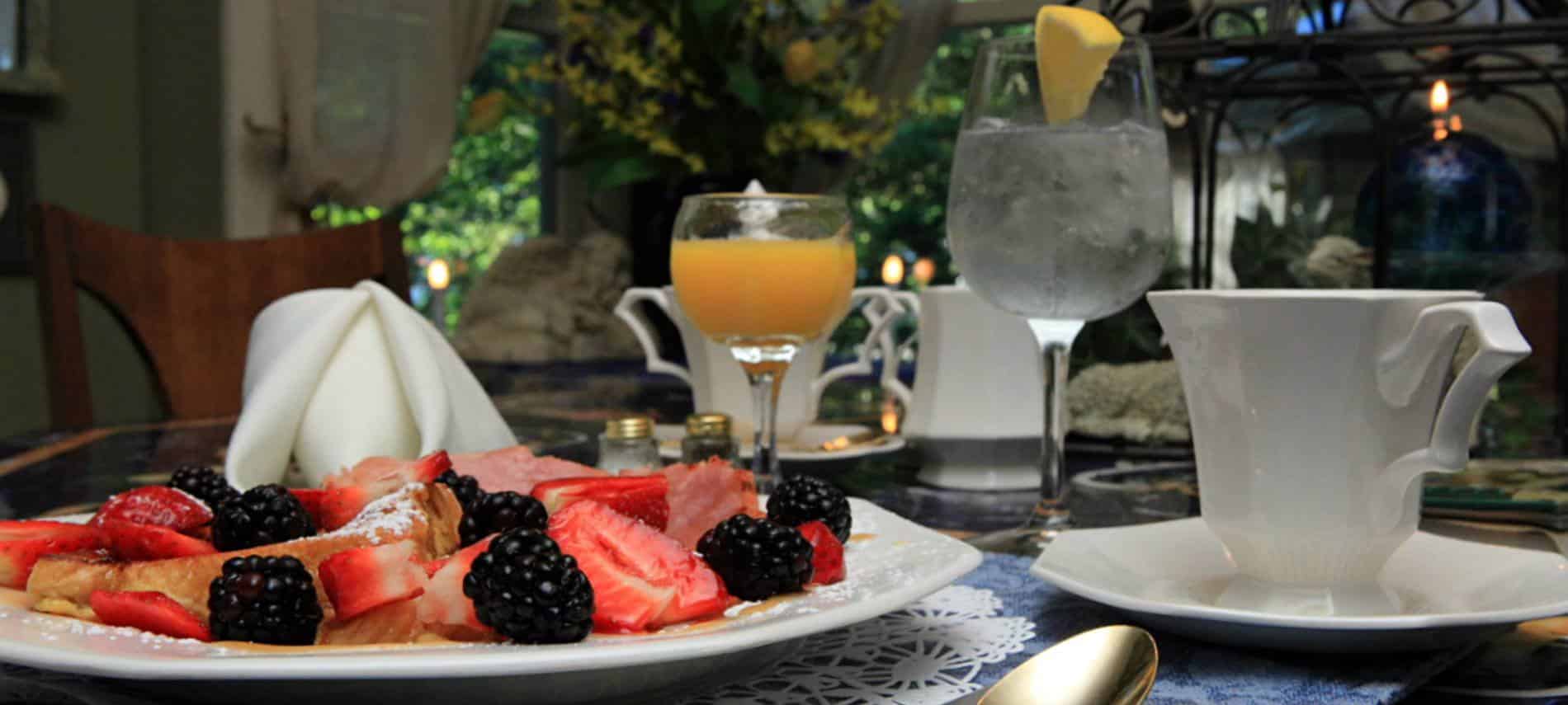 Table with white plate of French toast, fresh strawberries and blackberries, ham and white linen napkins, juice, water and coffee