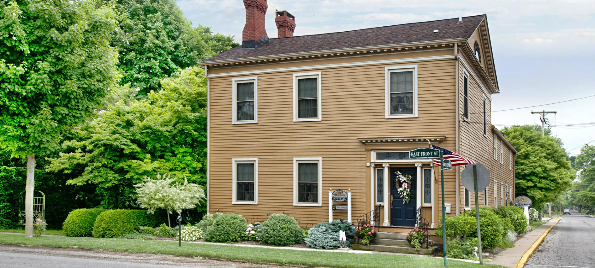 Exterior front view of B&B, tan siding, several windows, two brick chimneys surrounded by lush green trees and shrubs