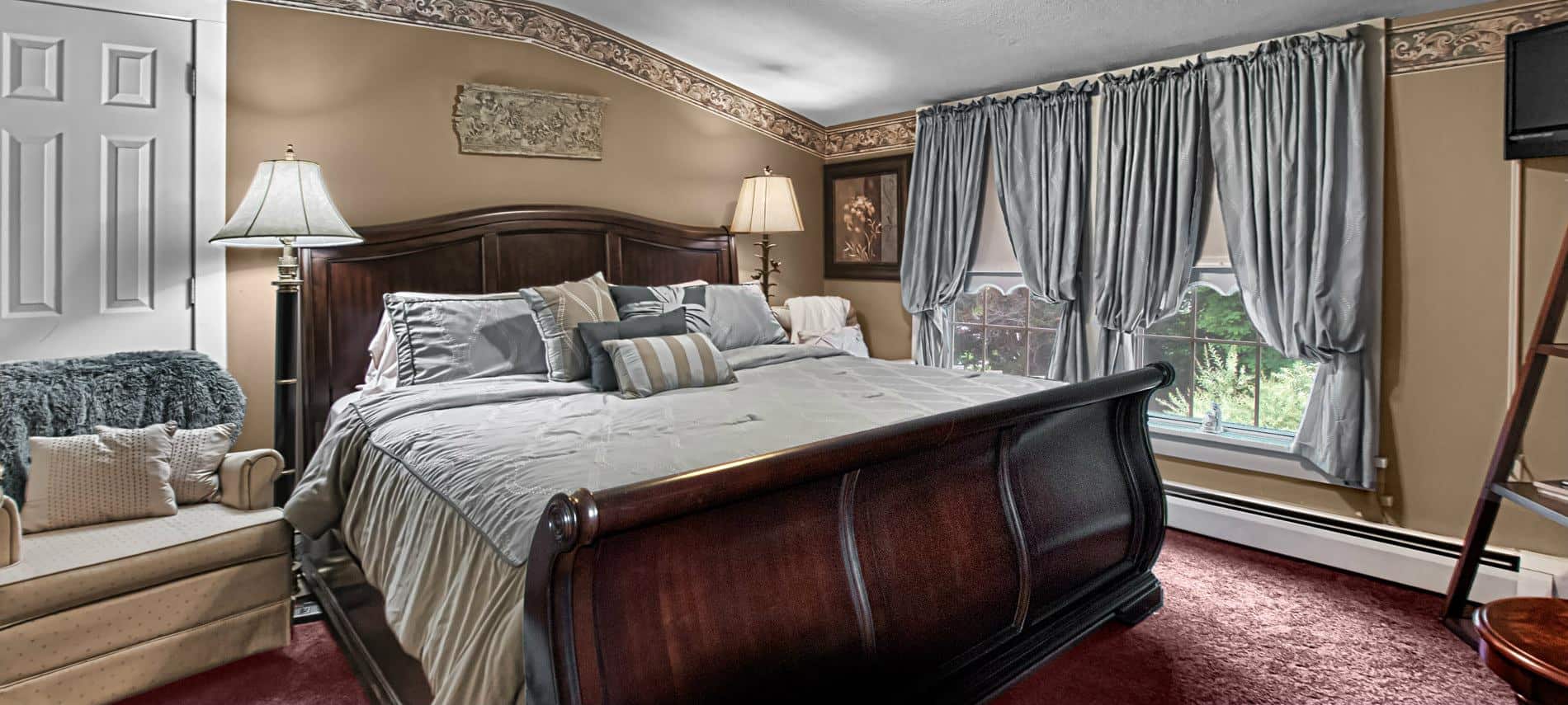 Elegant guest room with tan walls, burgundy carpet, double window, sleigh bed and upholstered chair