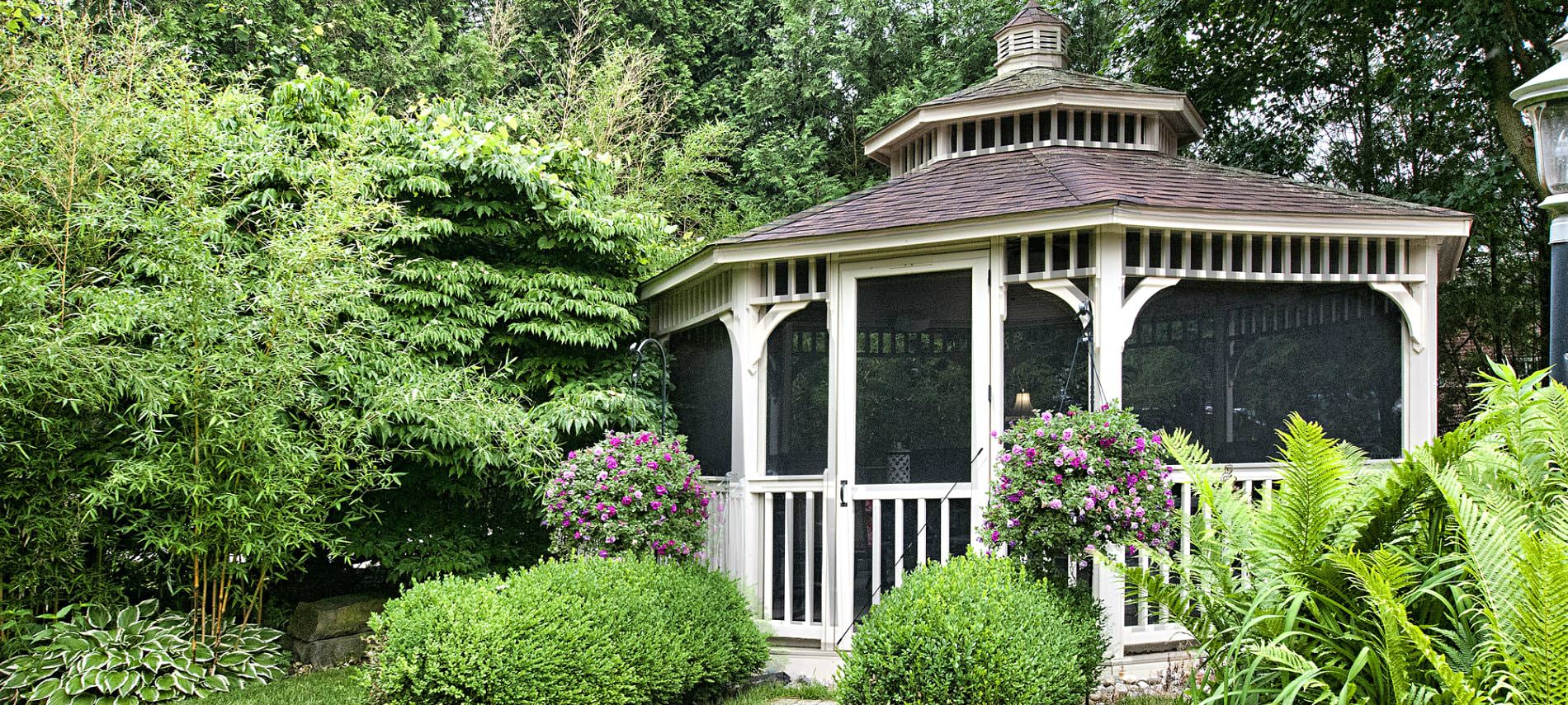 White screened-in gazebo with shingle roof and cupola surrounded by lush green trees and shrubs