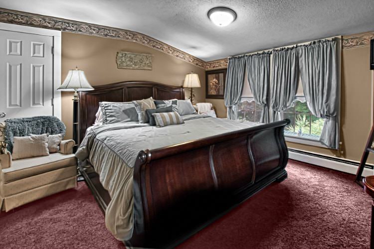 Elegant guest room with tan walls, burgundy carpet, double window, sleigh bed and upholstered chair