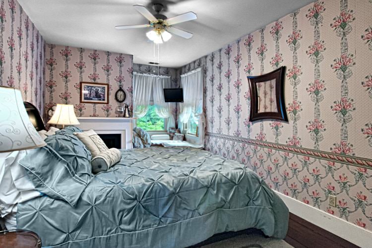 Pretty guest room with floral wallpaper, wood floors, wood bed with light blue shiny comforter, corner windows and fireplace