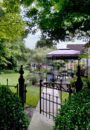 Side view of backyard with black metal fence and gate, patio with umbrella tables and lush landscaping