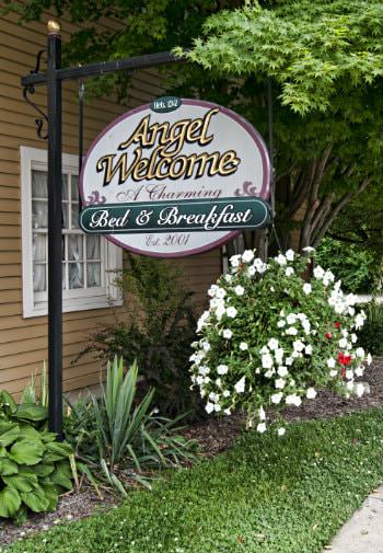 Exterior B&B black sign post with white oval hanging sign that says Angel Welcome A charming B&B established 2001