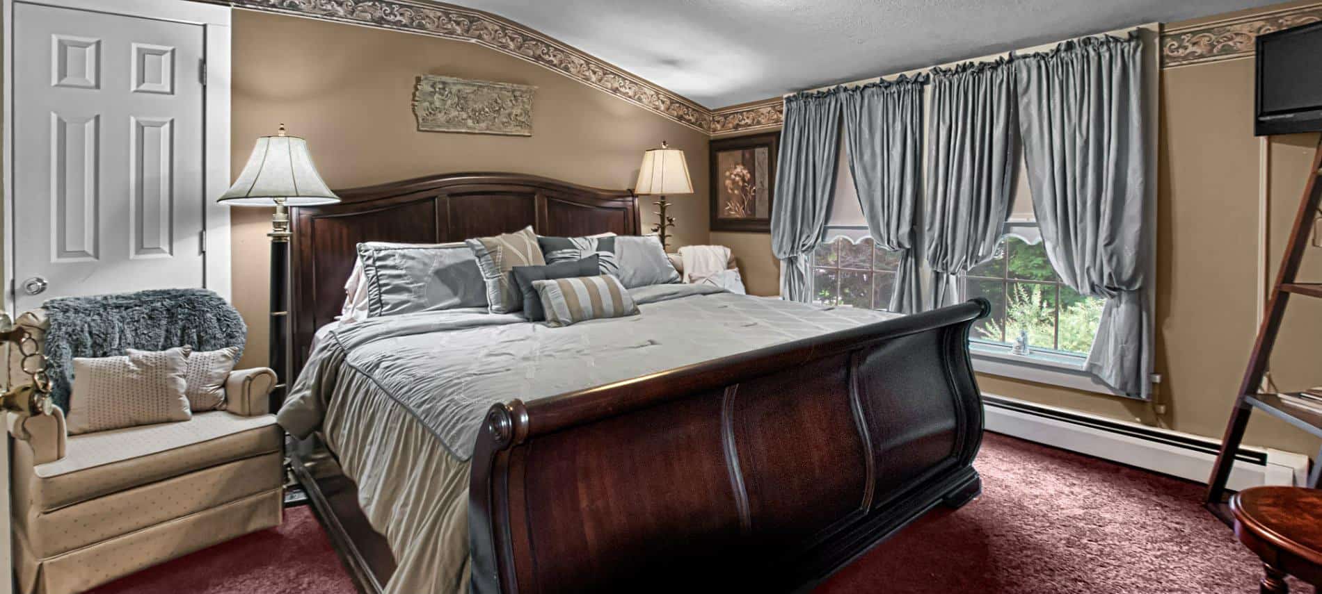 Elegant guest room with tan walls, burgundy carpet, wooden sleigh bed, large window and sitting chairs