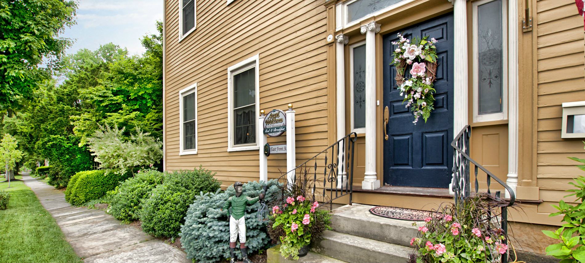 Front exterior of B&B, tan siding, steps up to a black door, lush green shrubs and trees